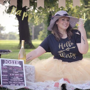 May include: A woman wearing a black t-shirt with gold glitter text that says "Hello Thirty" and a gold tulle skirt. She is holding a glass of champagne and wearing a black and white sun hat. A chalkboard sign with the text "JOSIE" and a list of facts about her is in the foreground.