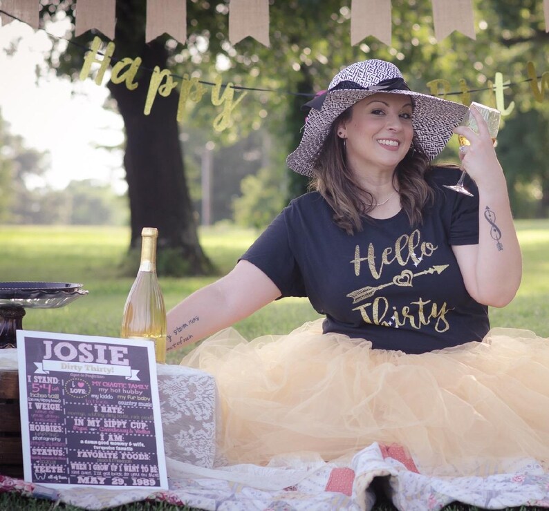 May include: A woman wearing a black t-shirt with gold glitter text that says "Hello Thirty" and a gold tulle skirt. She is holding a glass of champagne and wearing a black and white sun hat. A chalkboard sign with the text "JOSIE" and a list of facts about her is in the foreground.