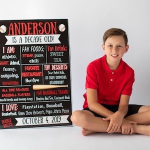May include: A black chalkboard sign with white text and red accents announcing "Anderson is a decade old!" The sign lists Anderson's favorite foods, drinks, desserts, and baseball teams. A young boy wearing a red polo shirt and black shorts sits in front of the sign.