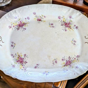 May include: An oval ceramic serving platter with a cream-colored base and a subtle crackle glaze finish. The platter features scattered floral clusters in shades of pink, purple, and yellow, with decorative gold-toned filigree accents on the edges.
