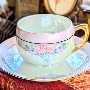 May include: A vintage teacup and saucer set. The cup and saucer are decorated with a pink and gold trim, floral designs, and iridescent accents. The teacup has a gold handle. The set is placed on a red book.