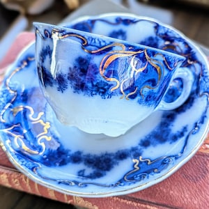 May include: A vintage teacup and saucer set. The cup and saucer are white with a blue floral design and gold trim. The cup has a handle and sits on a matching saucer. The set is likely made of porcelain.