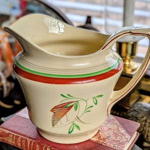 May include: Cream-colored ceramic pitcher with a wide spout and a curved handle. The pitcher features a red and green band around the middle and a bird and leaf design. The pitcher is sitting on a red book.