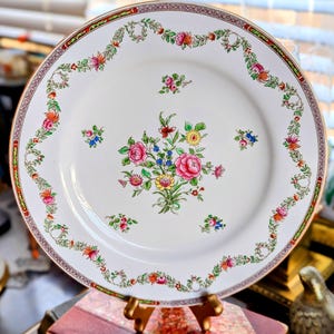 May include: A white ceramic plate with a floral design. The plate features a central bouquet of pink, yellow, and blue flowers, surrounded by a garland of flowers and leaves. The rim has a decorative border with red and gold accents.
