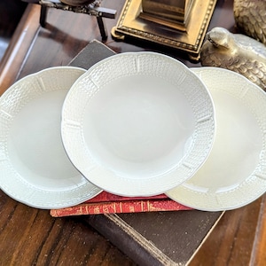 May include: Three white ceramic plates with textured rims and scalloped edges are stacked on a dark wooden surface. The plates have a simple, elegant design, suitable for serving food. The plates are a light cream color.