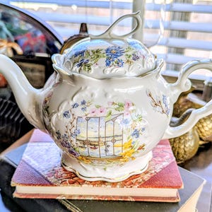 May include: White porcelain teapot with a lid, decorated with floral and landscape designs in blue, pink, and yellow. The teapot has a curved spout and handle, and sits on a stack of books.