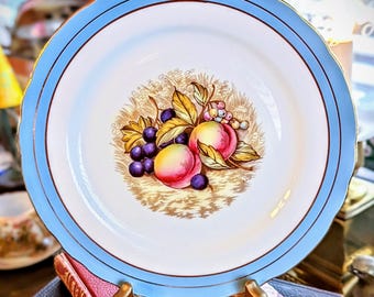 May include: A round ceramic plate with a scalloped edge, featuring a wide light blue border with a thin gold rim. The center displays a detailed illustration of peaches, grapes, and small berries nestled among green leaves on a tan background.