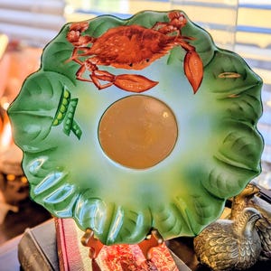 May include: A decorative plate with a crab and pea pod design. The plate is shaped like a large green leaf with a central circular indentation. The crab is orange and red, and the peas are green. The plate is supported by a gold-colored stand.