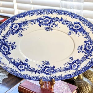 May include: An oval serving platter with a white center and a blue floral border. The blue floral design includes detailed flowers and foliage. The platter is made of ceramic and is likely vintage or antique.