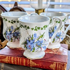 May include: Three white ceramic mugs with floral designs. Each mug features a blue and white floral pattern with green accents and gold trim. The mugs are placed on top of a stack of antique books.