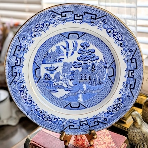 May include: A blue and white ceramic plate with a gold rim. The plate features a detailed Willow pattern design with a bridge, pagoda, and birds. The plate is displayed on a gold stand, showcasing its intricate details and classic style.