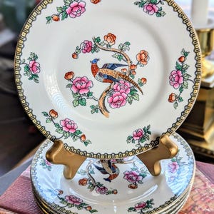 May include: A stack of vintage porcelain plates with a bird and floral design. The plates feature a cream-colored background with pink flowers, orange accents, and a gold rim. The bird is perched on a branch.