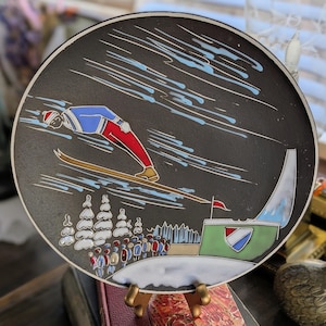 Puede incluir: Un plato decorativo negro con una escena de salto de esquí. El plato representa a un esquiador en el aire, con un atuendo rojo, azul y blanco, con un trampolín y espectadores en el fondo. El plato tiene un borde blanco.