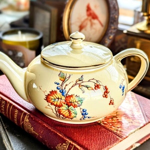 May include: Cream-colored vintage teapot with a lid, featuring a hand-painted design of colorful leaves and berries. The teapot has a gold trim and sits atop a stack of antique books with red covers. A classic piece for tea lovers.