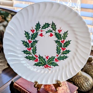 May include: White ceramic plate with a swirled rim design. The plate features a central wreath design of green holly leaves and red berries, perfect for holiday or seasonal decor. The plate is displayed on a gold-colored stand.