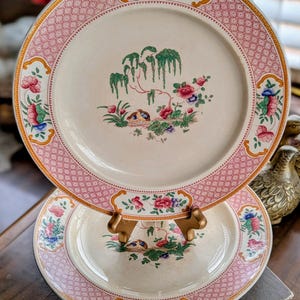 May include: Two antique porcelain plates with a pink and cream color scheme. The plates feature a floral and tree design in the center, with a pink patterned border and decorative floral accents. The plates are displayed on a gold-colored stand.