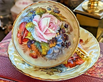 Può includere: Servizio da tè vintage con tazza e piattino color crema, bordati d'oro. La tazza presenta una grande rosa rosa circondata da altri fiori colorati. Decorazione floreale.
