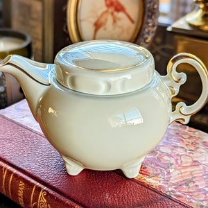 May include: A small, cream-colored porcelain teapot with a lid, featuring gold trim on the handle and rim. The teapot has a rounded body, a curved spout, and sits on three small feet. The background includes a book and framed artwork.