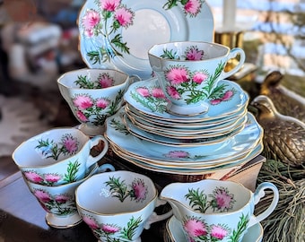 May include: A porcelain tea set featuring a light blue base color with hand-painted pink thistle flowers and green leaves. The set includes multiple teacups, saucers, a creamer, and a plate, all finished with delicate gold trim on the scalloped edges.