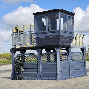 May include: A gray and yellow toy military base with a watchtower and fencing. A soldier figure in green and black camouflage stands in front of the base. The sky is blue with white clouds.