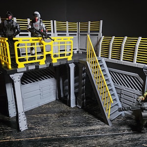 May include: A grey and yellow playset with action figures. The playset features a platform with yellow railings, a staircase, and a detailed structure. Two figures stand on the platform, while another figure stands below with a large weapon.
