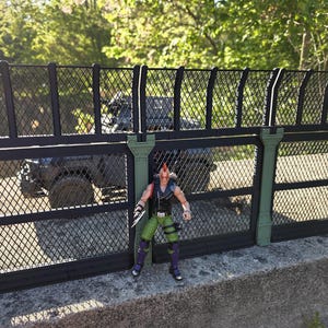 Può includere: Una recinzione metallica nera e verde con un motivo a maglia diamantata. Una statuetta con pantaloni verdi, stivali viola e un'acconciatura a cresta rossa è in piedi di fronte alla recinzione. Un veicolo nero è visibile dietro la recinzione.