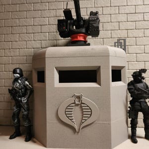 Op de afbeelding: Een grijs, rechthoekig Cobra-commandocentrum met een gemonteerd kanon, geflankeerd door twee zwarte actiefiguren. Het commandocentrum heeft een Cobra-logo en rechthoekige openingen. De figuren dragen tactische uitrusting.