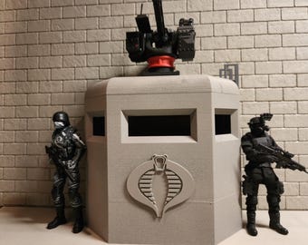 Può includere: Un centro di comando Cobra grigio rettangolare con una mitragliatrice montata, affiancato da due action figure nere. Il centro di comando presenta un logo Cobra e aperture rettangolari. Le figure indossano equipaggiamento tattico.