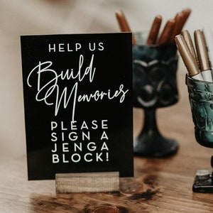 Puede incluir: Un letrero negro con texto blanco que dice "Help us Build Memories. Please sign a Jenga block!" con un soporte de madera. Un jarrón de vidrio con marcadores dorados y blancos.