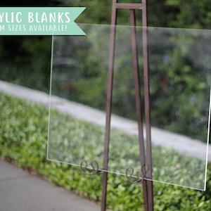 May include: A clear acrylic blank sign on a metal easel stand. The sign is on a pavement in front of a green bush. The sign reads "Acrylic Blanks Custom Sizes Available!"