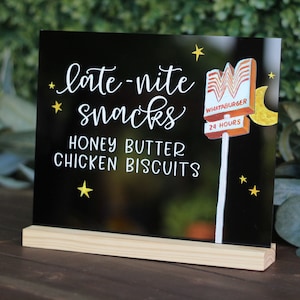 May include: Black sign with white text that reads "late-nite snacks honey butter chicken biscuits". A stylized illustration of a Whataburger sign with a yellow moon and stars is on the right side of the sign. The sign is on a wooden stand.