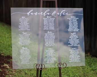 May include: A clear acrylic wedding seating chart with the names "Daniela & Alec" at the top. The chart is divided into nine sections, each labeled with a number from one to nine. Each section lists the names of guests assigned to a specific table.