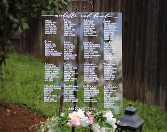 May include: A clear acrylic wedding seating chart with white lettering. The chart is divided into 16 sections, each labeled with a number and the names of guests. The chart is titled "Michelle and Berich".