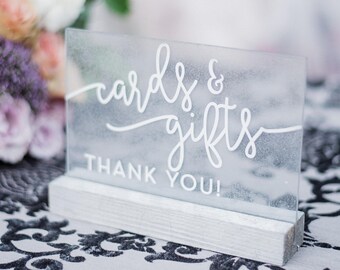 May include: A frosted glass sign with white lettering that reads "cards & gifts thank you!" The sign is sitting on a white wooden base and is on a black and white patterned surface.