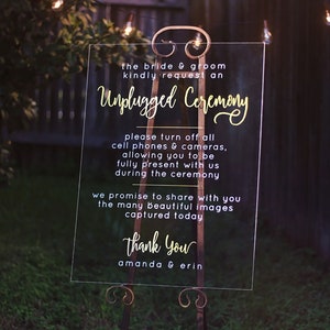 May include: A clear acrylic sign with gold lettering that reads "The bride & groom kindly request an Unplugged Ceremony. Please turn off all cell phones & cameras, allowing you to be fully present with us during the ceremony. We promise to share with you the many beautiful images captured today. Thank you, Amanda & Erin."