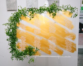 May include: A clear acrylic wedding seating chart with a gold background and white brushstrokes. The chart is decorated with greenery and lists the names of guests.