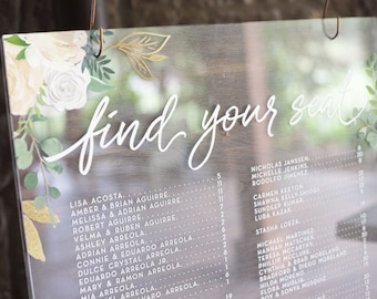 May include: A clear acrylic sign with a floral design and the text "find your seat" printed in white. The sign lists names and table numbers for a wedding reception.