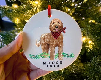 May include: A round white ceramic ornament featuring a brown curly-haired dog wearing a red and black buffalo plaid bandana. The dog stands above green pine branches. Below the dog, the text reads MOOSE 2018 in black capital letters.