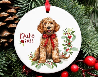 Puede incluir: Un adorno redondo de cerámica blanca colgado de una cinta roja, con una ilustración en acuarela de un perro doodle marrón dorado con un lazo rojo, rodeado de hojas de acebo verdes y bayas rojas, con el texto Duke y 2025.
