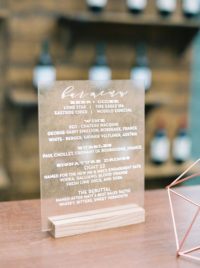 May include: A clear acrylic sign with a wooden base lists a bar menu with beer, cider, wine, bubbles, and signature drinks. The sign reads "Carmenn" at the top and lists the drinks with their names and origins.