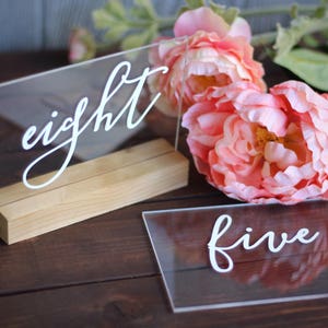 May include: Two clear acrylic table numbers with the words "eight" and "five" written in white calligraphy. The numbers are on wooden stands and are surrounded by pink flowers.