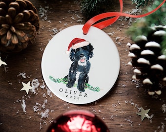 Könnte beinhalten: Ein runder, weißer Keramik-Weihnachtsanhänger mit einem Aquarellbild eines schwarzen Hundes mit roter Weihnachtsmütze, der auf grünen Tannenzweigen sitzt. Der Text OLIVER 2023 ist in Schwarz unter dem Hund aufgedruckt. Rotes Band befestigt.