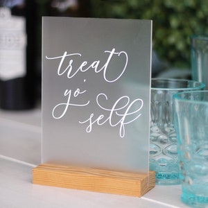 May include: A frosted acrylic sign with a wooden base that reads "treat yo self" in white cursive lettering.