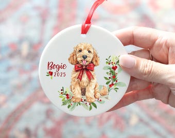 Può includere: Un ornamento rotondo in ceramica bianca con un'illustrazione ad acquerello di un cane color oro che indossa un fiocco rosso. Il testo Bogie 2025 è stampato in rosso accanto al cane, circondato da foglie di agrifoglio verdi e bacche rosse.
