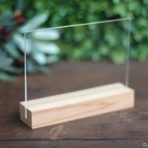 May include: A clear acrylic sign holder with a natural wood base. The sign holder is standing on a wooden surface.