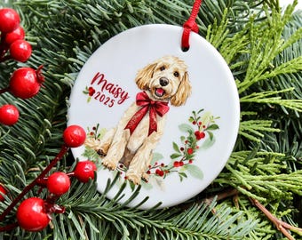 Può includere: Un ornamento rotondo in ceramica bianca con un'illustrazione ad acquerello di un cane color oro che indossa un fiocco rosso. Il testo "Maisy" e "2025" è stampato in rosso, accompagnato da una ghirlanda festiva di foglie verdi e bacche rosse.