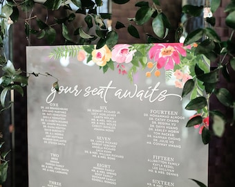 May include: A gray acrylic seating chart with white text and floral illustrations. The text reads "your seat awaits" and lists names and table numbers.