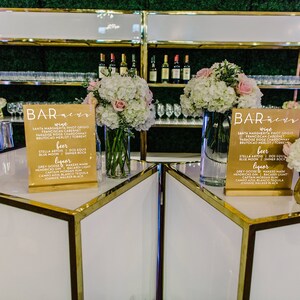 May include: Two white bar tables with gold trim and a gold shelf behind them. Each table has a sign that says "Bar Menu" with a list of drinks. The tables are decorated with white and pink flowers.