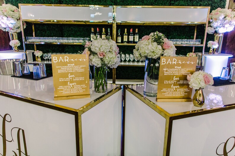 May include: Two white bar tables with gold trim and a gold shelf behind them. Each table has a sign that says "Bar Menu" with a list of drinks. The tables are decorated with white and pink flowers.