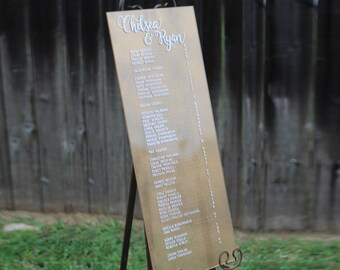 May include: A gold-colored wedding seating chart with white lettering. The chart is on an easel and lists the names of wedding guests.
